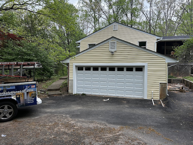 Tan two-story garage with white door, parked truck on asphalt driveway surrounded by trees